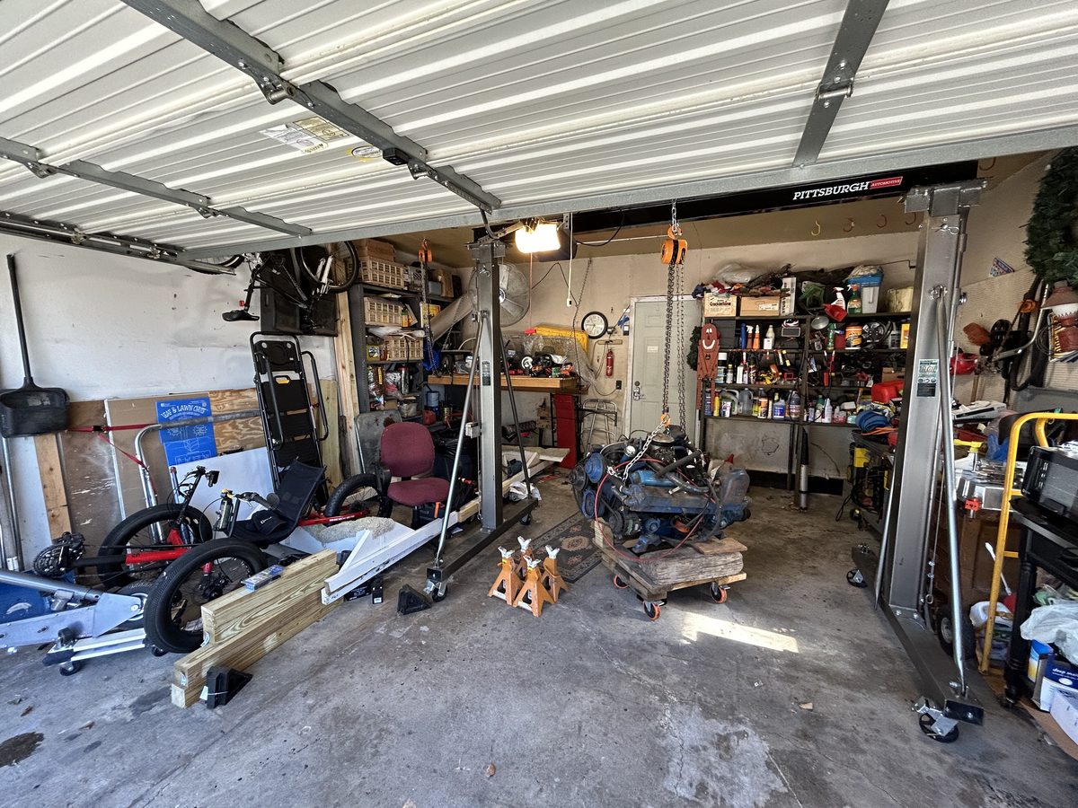 Home garage with a small-block V8 hanging on an engine hoist and parts on the shelves. Photo: TruckMatt, CC0, via Wikimedia Commons