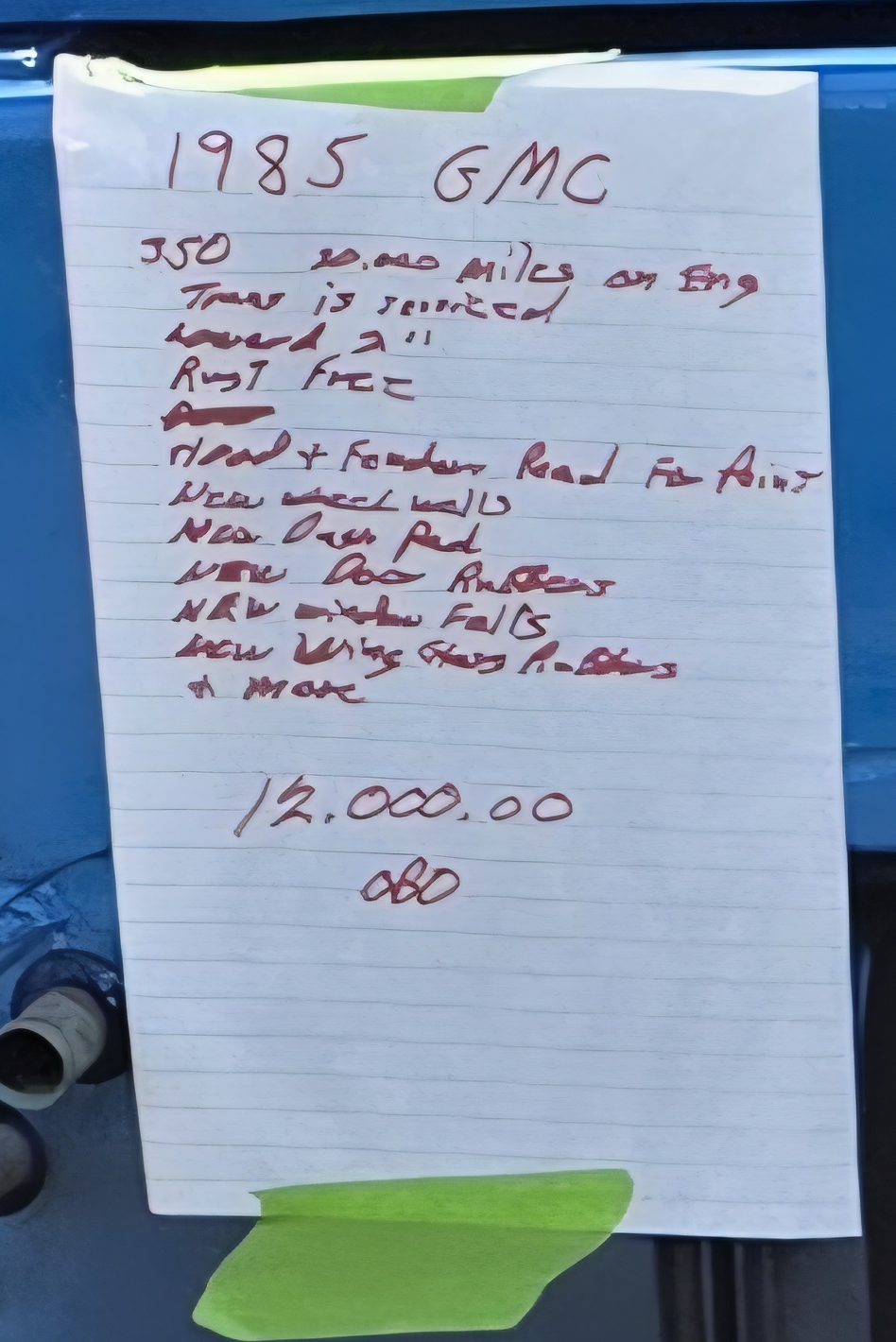 Close-up of a handwritten for-sale sheet taped to a blue truck: 1985 GMC, 350, miles on engine, trans serviced, lowered 2 inches, rust free, hood and fenders ready for paint, list of new rubber and trim parts, 12,000.00 OBO