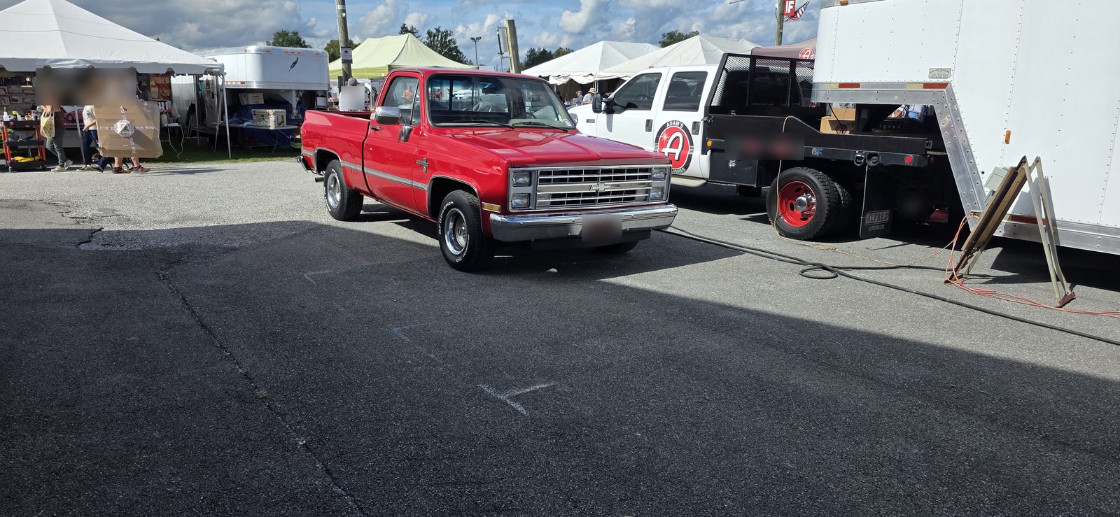 A clean red 1981 to 1987 Chevrolet C10 short bed pickup parked on the asphalt at Fall Carlisle, next to a white parts-hauler truck and a box trailer, with vendor tents in the background