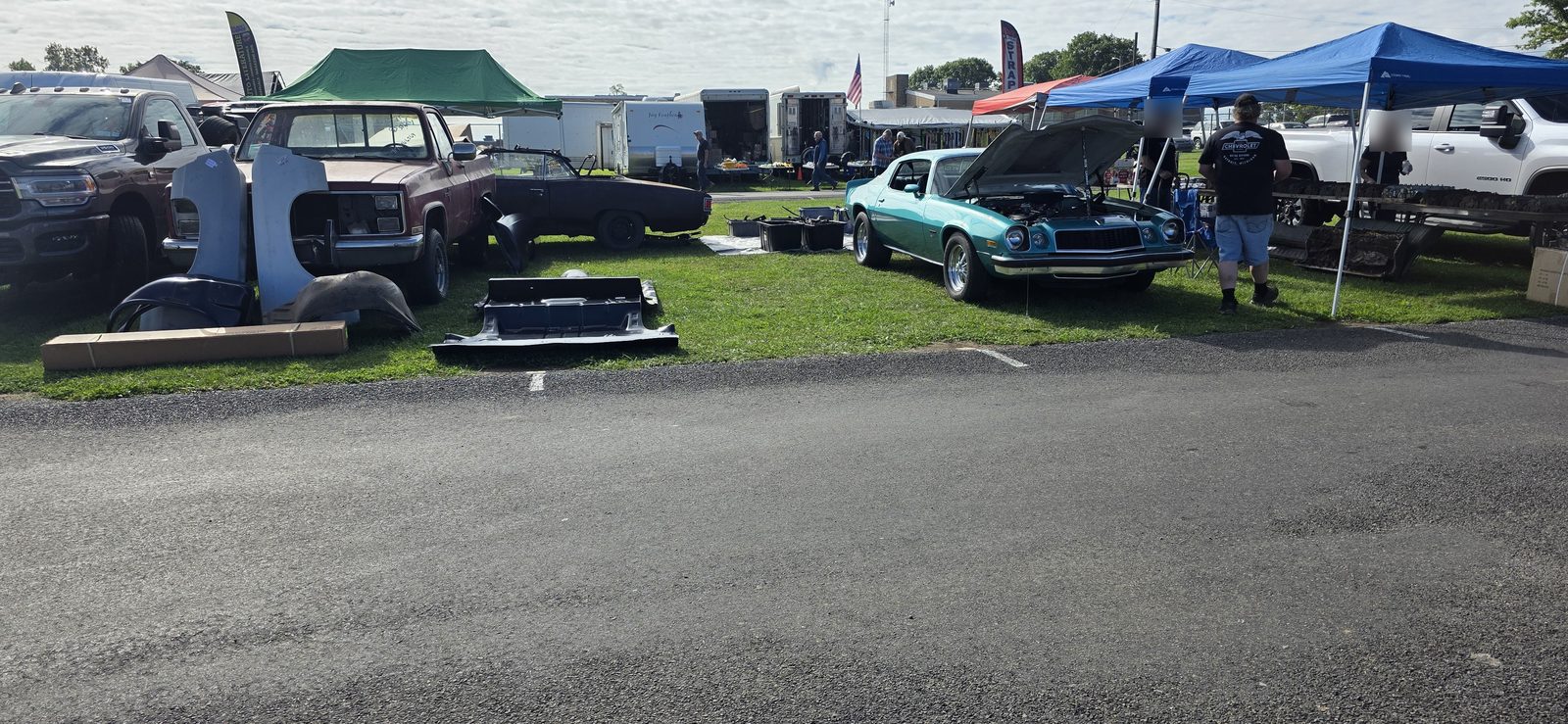 A row of vendor spaces on the grass at Fall Carlisle: reproduction fenders and inner fenders standing in front of a squarebody pickup, a stamped panel laid flat, a black convertible project car, a teal second-generation Camaro with the hood up, and blue vendor tents with used parts on racks